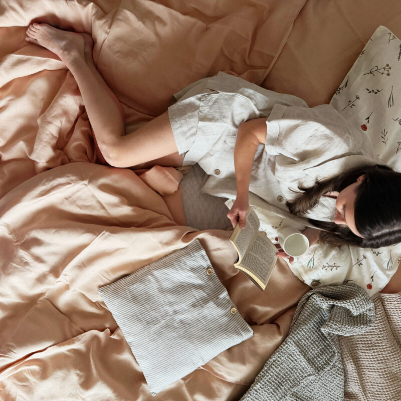 Záber z nadhľadu na útulnú posteľ s marhuľovými ľanovými obliečkami Adela, na ktorej leží žena v ľanovom pyžame a číta knihu.