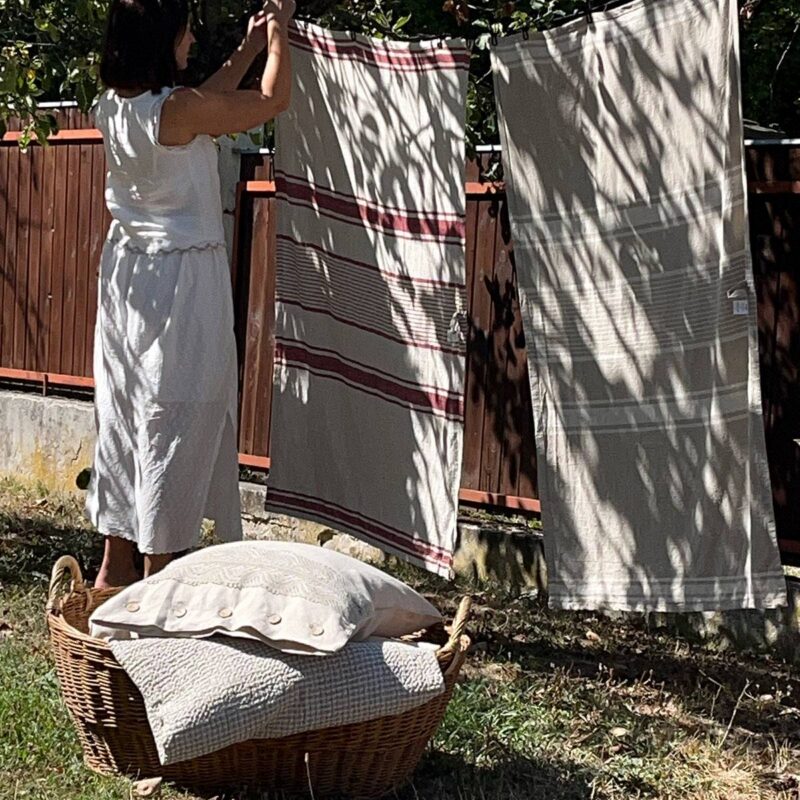 Lněná pruhovaná osuška červená a přírodní a žena, která věší lněné pruhované osušky na šňůru na zahradě, v popředí proutěný koš s lněným povlečením